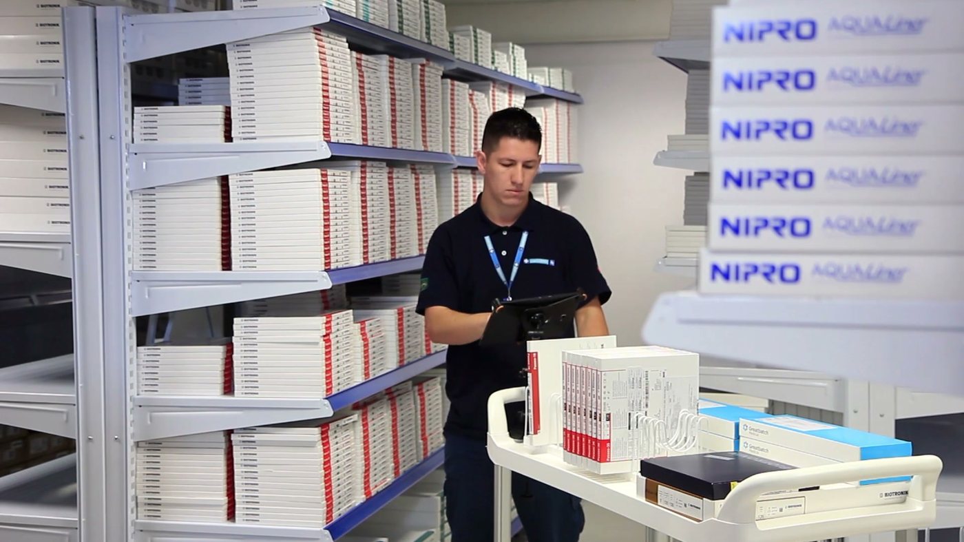 Centro de distribuição da Discomed: prateleiras com caixas de produtos Nipro e colaborador conferindo itens em tablet junto ao carrinho de separação.